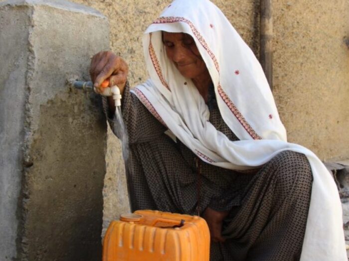 an afghan woman smiling as she collects water from a tap