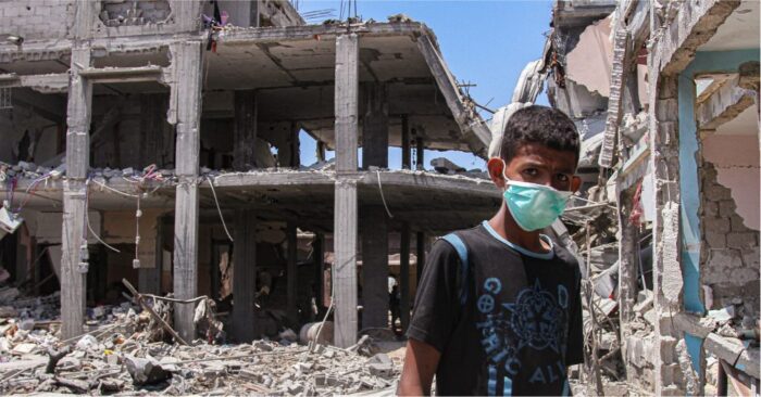 child with a blue face mask standing infront of destroyed buildings