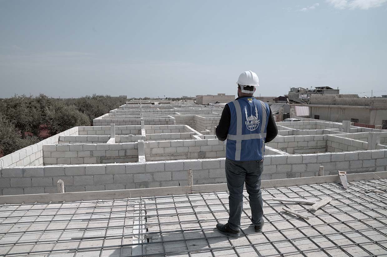 man wearing islamic relief jacket and safety helmet, standing on building overlooking construction of new homes being built syria earthquake