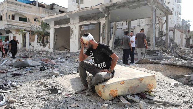 an injured man in gaza sitting down with his hand to his head, sat amongst debris from buildings destroyed from airstrikes