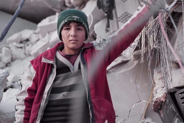 islamic relief boy in green hat and red coat standing next to broken buildings turkiye syria earthquake appeal