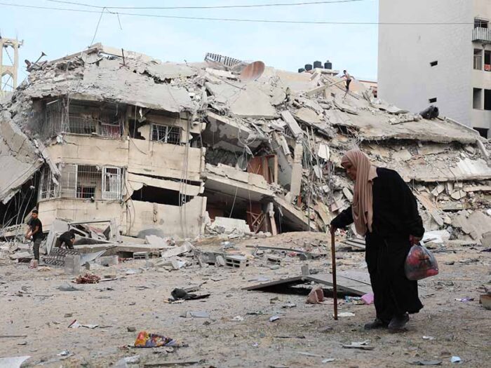 elderly woman walking in gaza alongside collapsed buildings destroyed from recent airstrikes