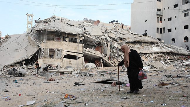 elderly woman walking in gaza alongside collapsed buildings destroyed from recent airstrikes