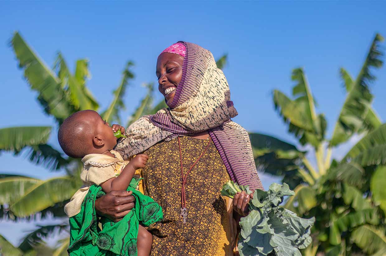 a women holding her baby and smiling at the baby with a handful of vegetable leaves in one hand funding futures types of sadaqah