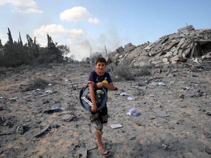 a young boy holding his belongings as he flees from destroyed buildings in gaza