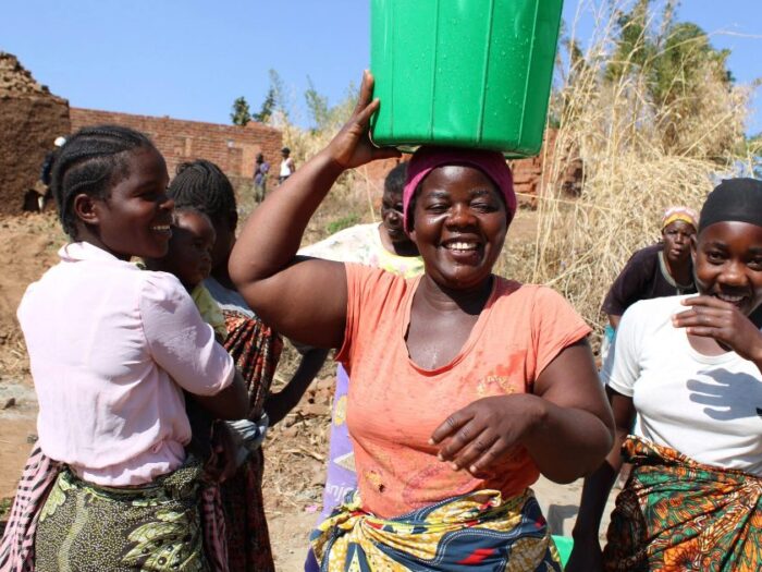 a woman in malawi smiling with a water bucket over her head and surrounded by other women looking joyous