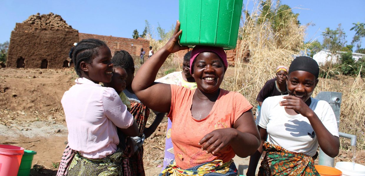 a woman in malawi smiling with a water bucket over her head and surrounded by other women looking joyous