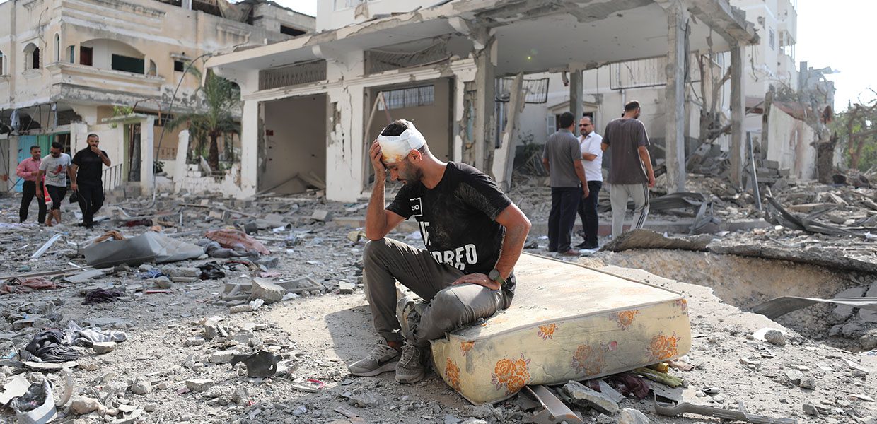 an injured man in gaza sitting down with his hand to his head, sat amongst debris from buildings destroyed from airstrikes