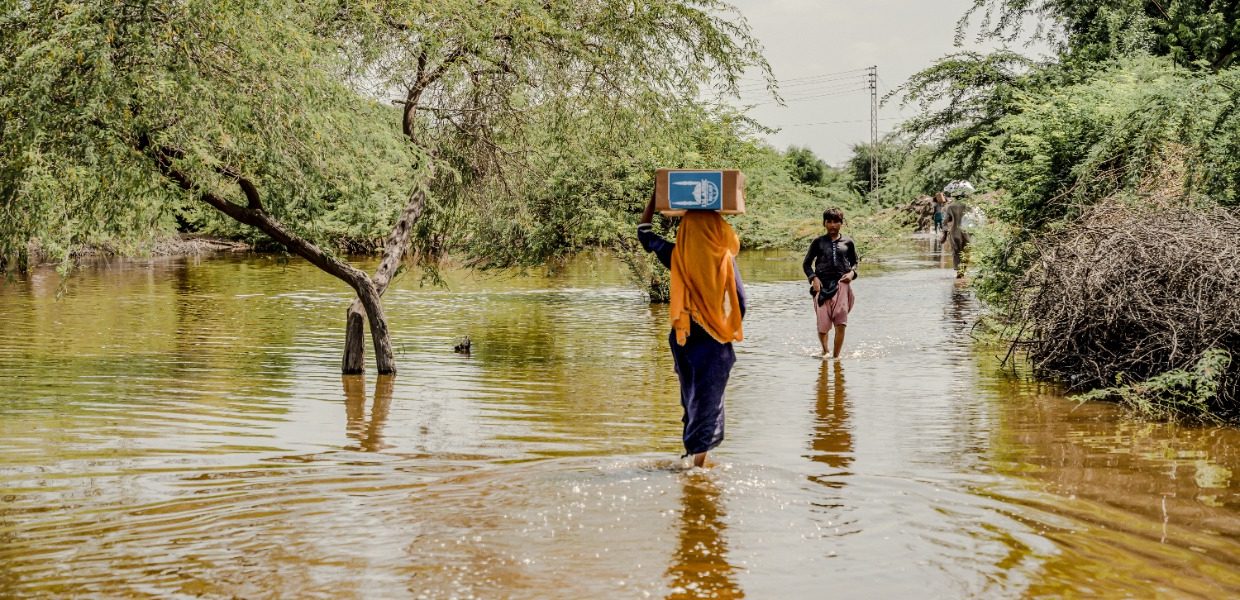 a woman in a orange hijab carrying an islamic relief box over her head while walking through a flooded road