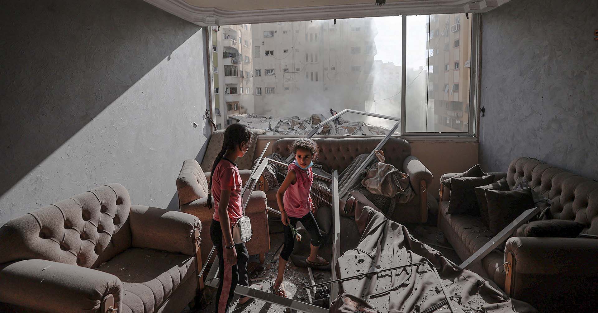 two children in gaza palestine standing in a room with a broken furniture and windows, with smoke filling the air outside window from debris and airstrikes