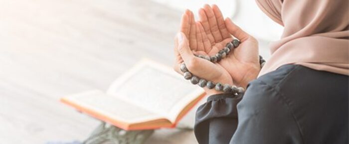 women with hands open in front of her for dua with prayer beads (tasbih) in her hands and an open quran placed in front of her prayer timetables
