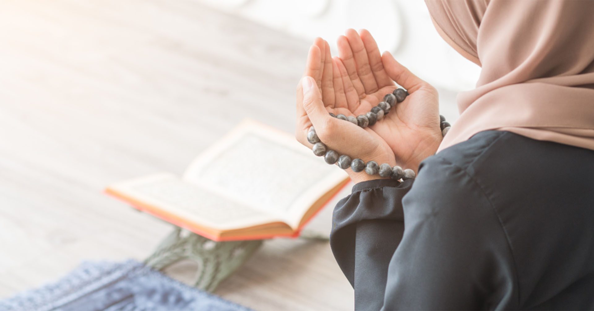 women with hands open in front of her for dua with prayer beads (tasbih) in her hands and an open quran placed in front of her prayer timetables high wycombe