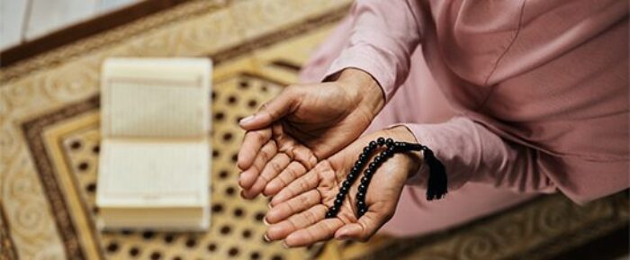women with hands open in front of her for dua with prayer beads (tasbih) in her hands and an open quran placed in front of her prayer timetables