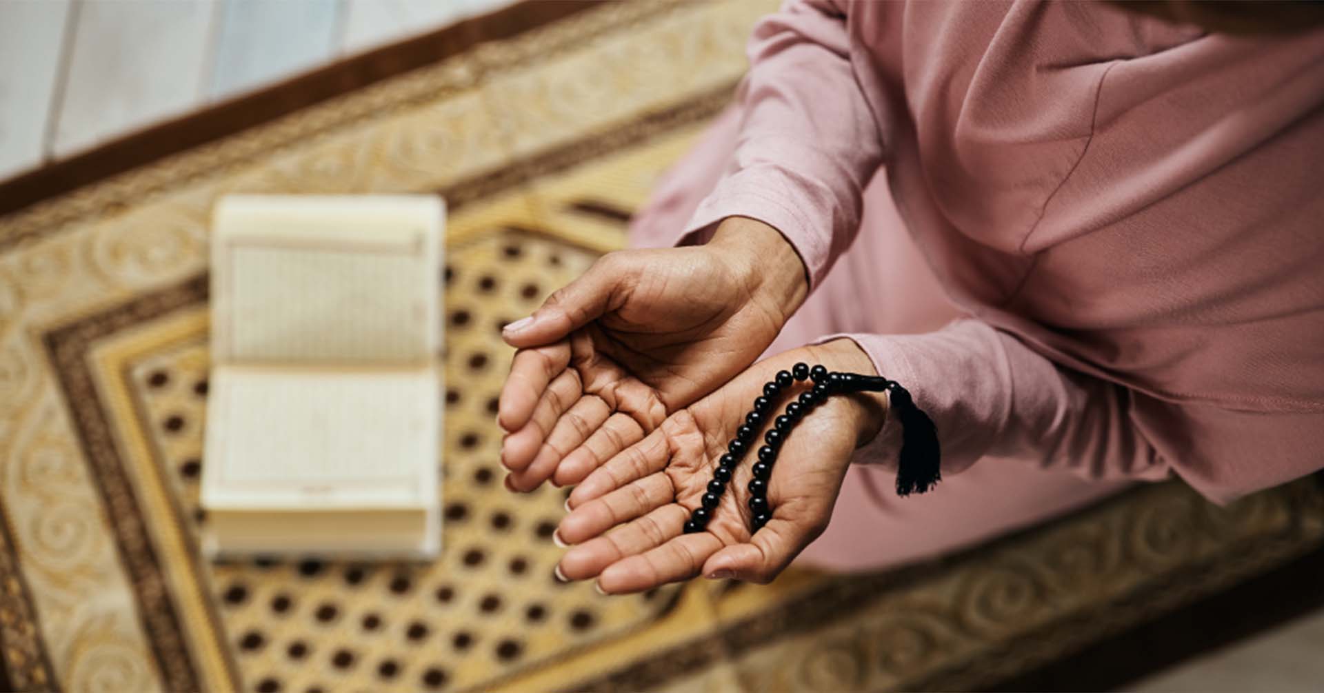 women with hands open in front of her for dua with prayer beads (tasbih) in her hands and an open quran placed in front of her prayer timetables bolton