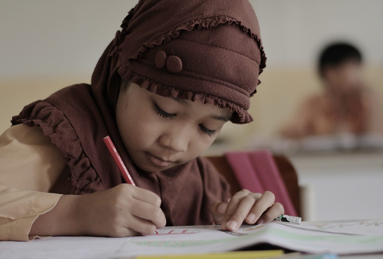 a young girl from indonesia, wearing a brown hijab is sat on a desk writing onto a book