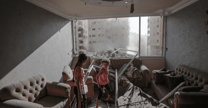 two children in gaza palestine standing in a room with a broken furniture and windows, with smoke filling the air outside window from debris and airstrikes