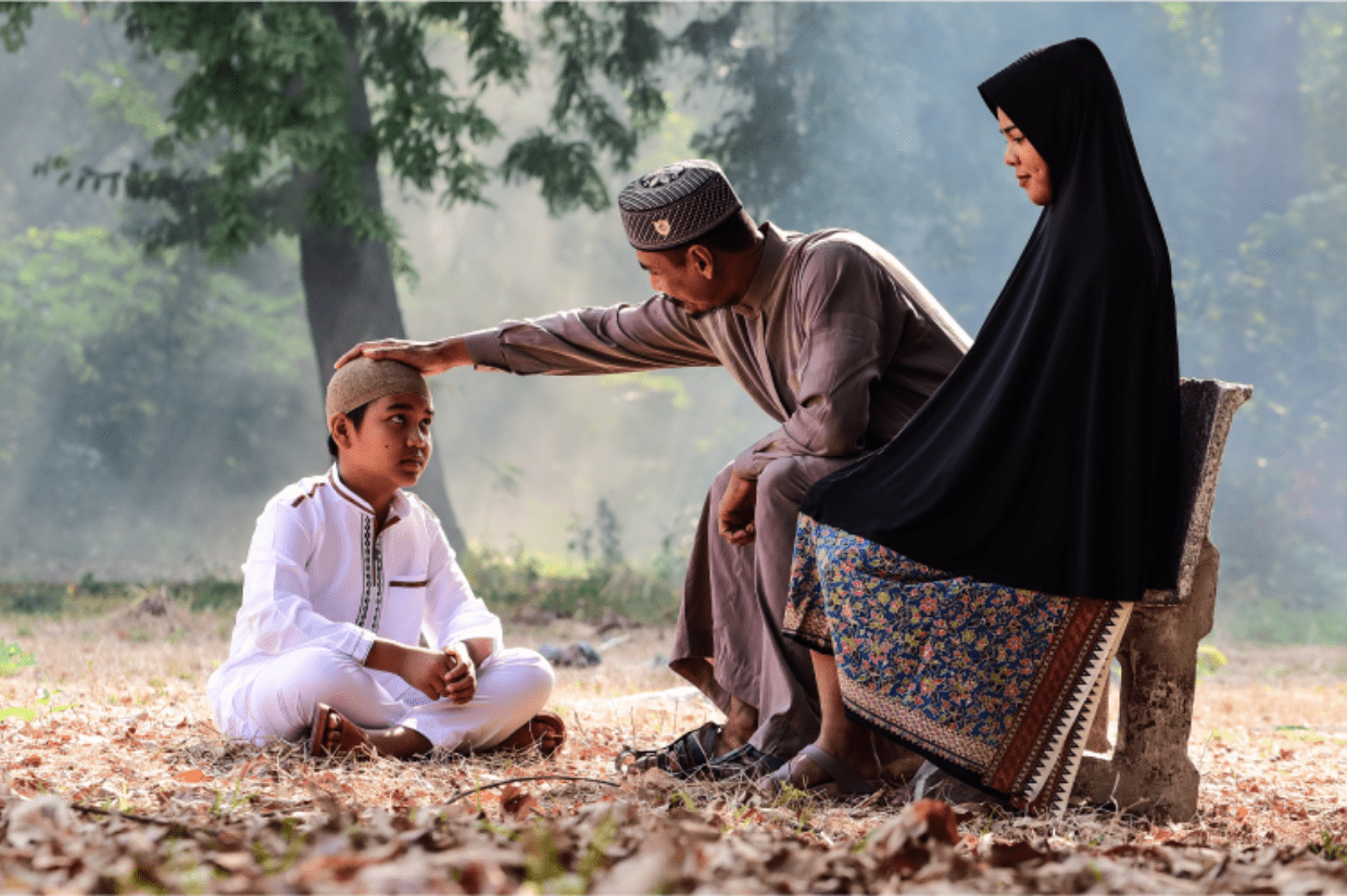 young boy sat on the grass and his head being patted by his father, with his mother sitting nearby sadaqah for parents