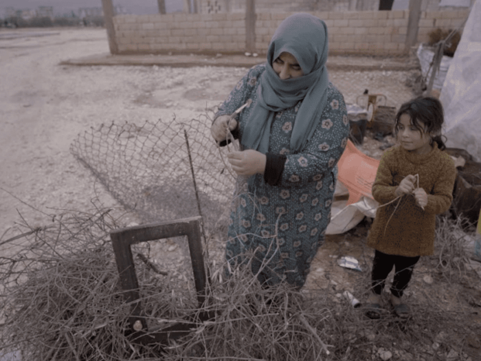 a syrian mother and daughter in a camp gathering twigs and bits of wood